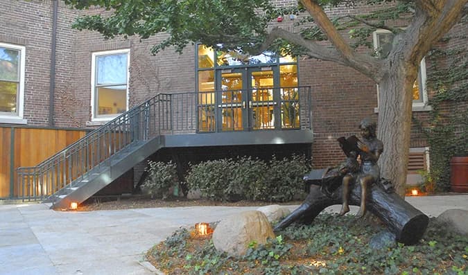 School courtyard with a brick facade, glazed entrance, mature tree, and reading sculpture