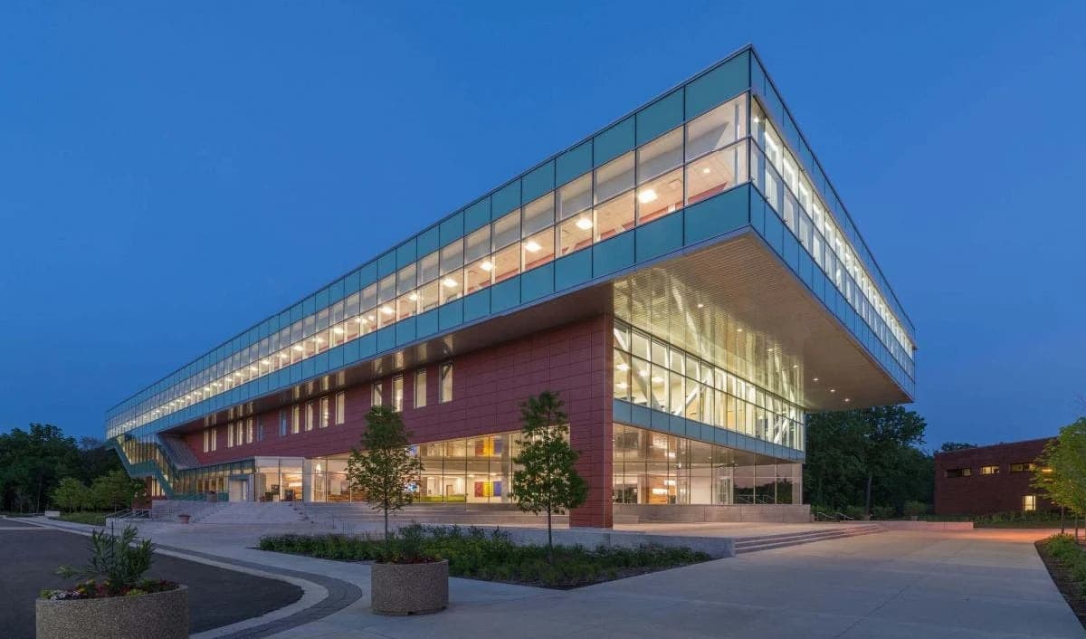 Academic building at dusk with illuminated glass walls, a cantilevered upper floor, and a landscaped entry plaza
